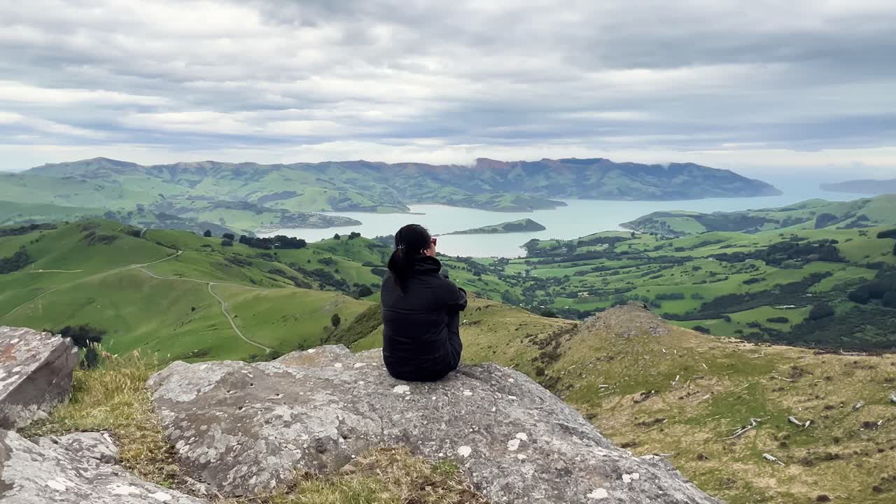 Person sitting on rock admiring Akaroa Peninsula, New Zealand landscape