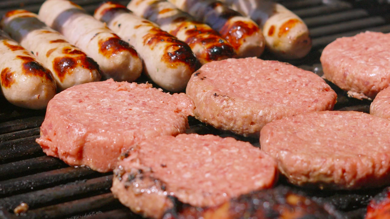 Ripresa al rallentatore di hamburger di manzo freschi e crudi che iniziano a cuocere su una griglia calda per barbecue sotto la luce intensa del sole. Gustoso cibo estivo. Salsicce sullo sfondo. 4K.