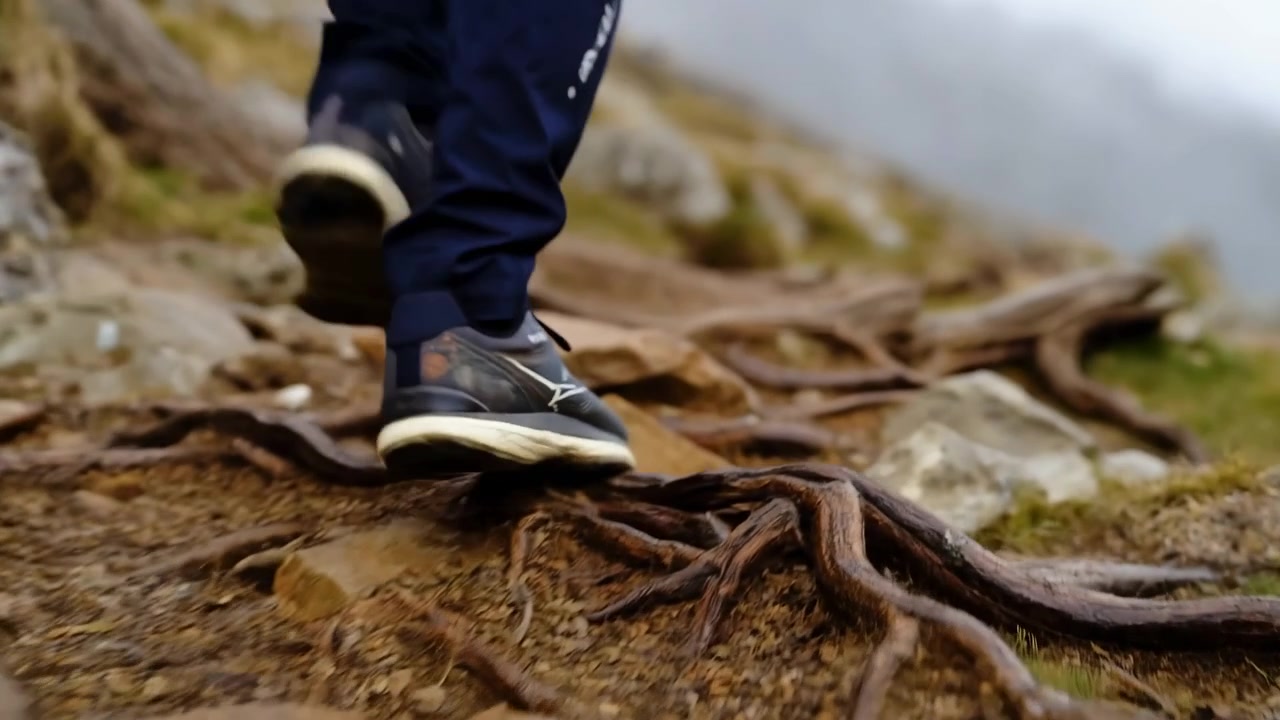 Vista a livello del suolo delle gambe inferiori, con pantaloni sportivi scuri e scarpe da ginnastica ammortizzate con suole chiare, che attraversano un terreno accidentato (rocce, radici, terra) su un sentiero, sotto una luce fioca e nuvolosa, in un contesto di escursionismo o trail running