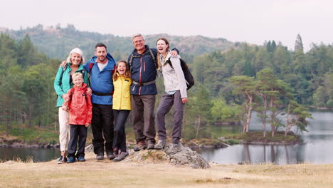 Familia-De-Varias-Generaciones-De-Pie-Sobre-Las-Rocas-Junto-A-Un-Lago-En-El-Campo-Mirando-A-La-Cámara,-Lake-District,-Reino-Unido