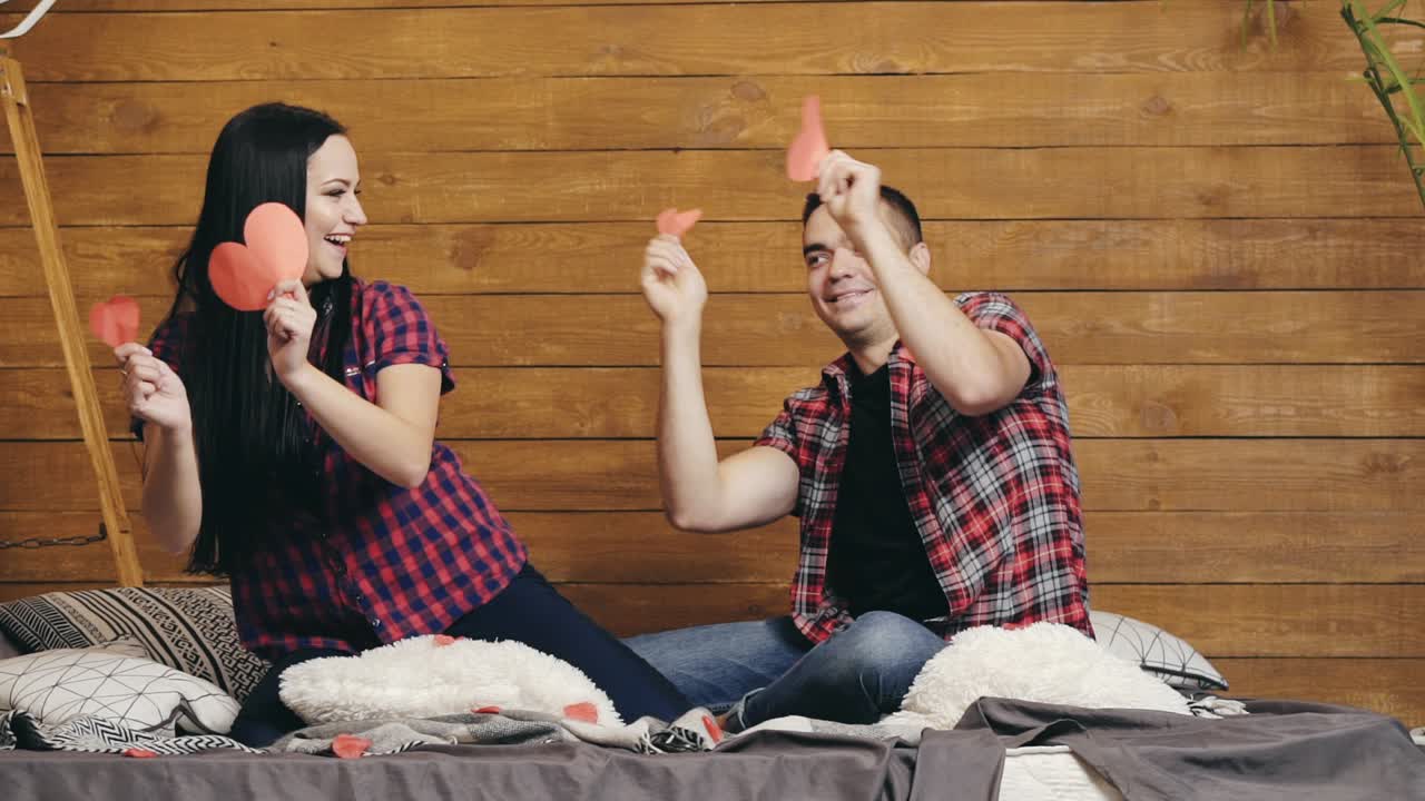 funny couple are sitting on the bed and playing with red paper hearts in their hands on Valentine's Day