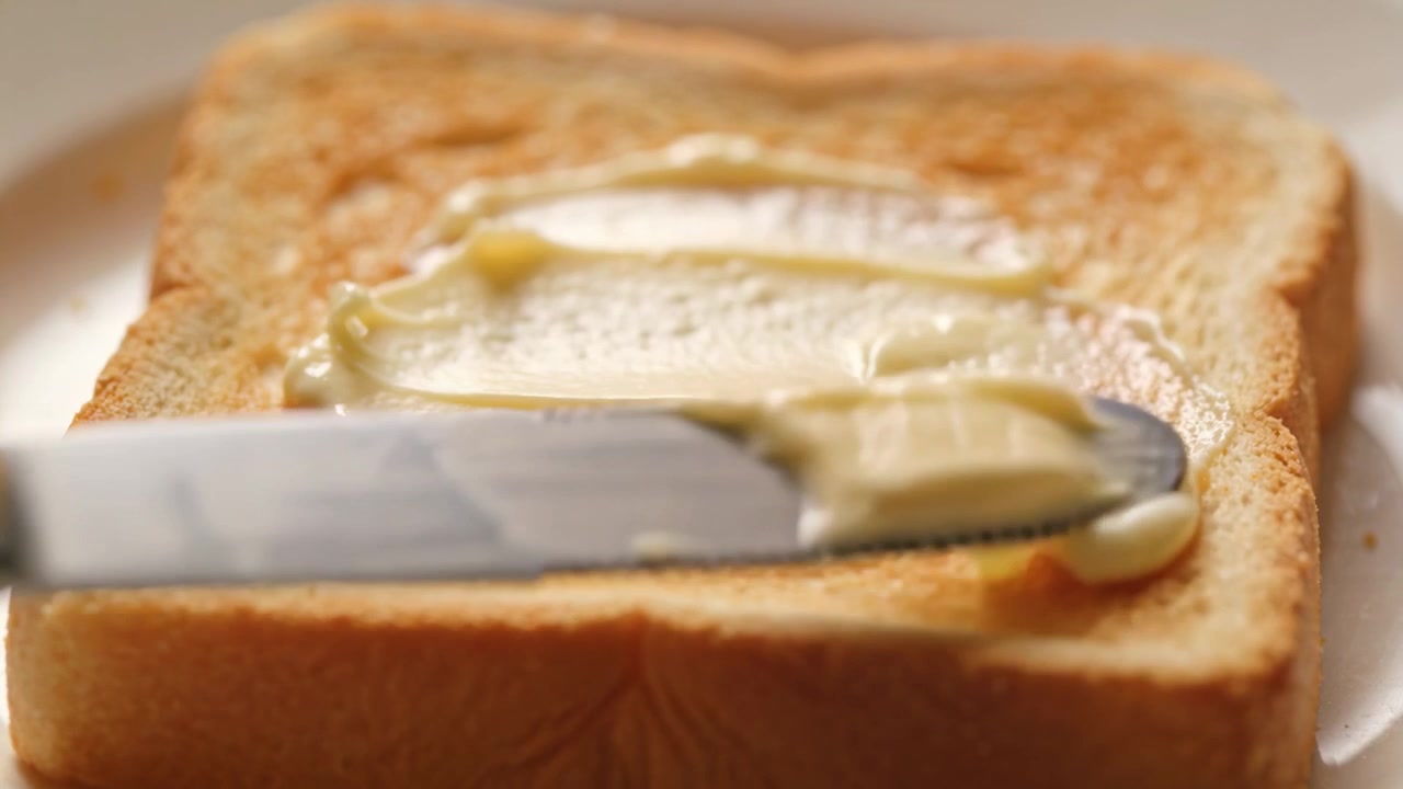Spreading person's hand dragging serrated knife across butter blob on toast on plate for breakfast
