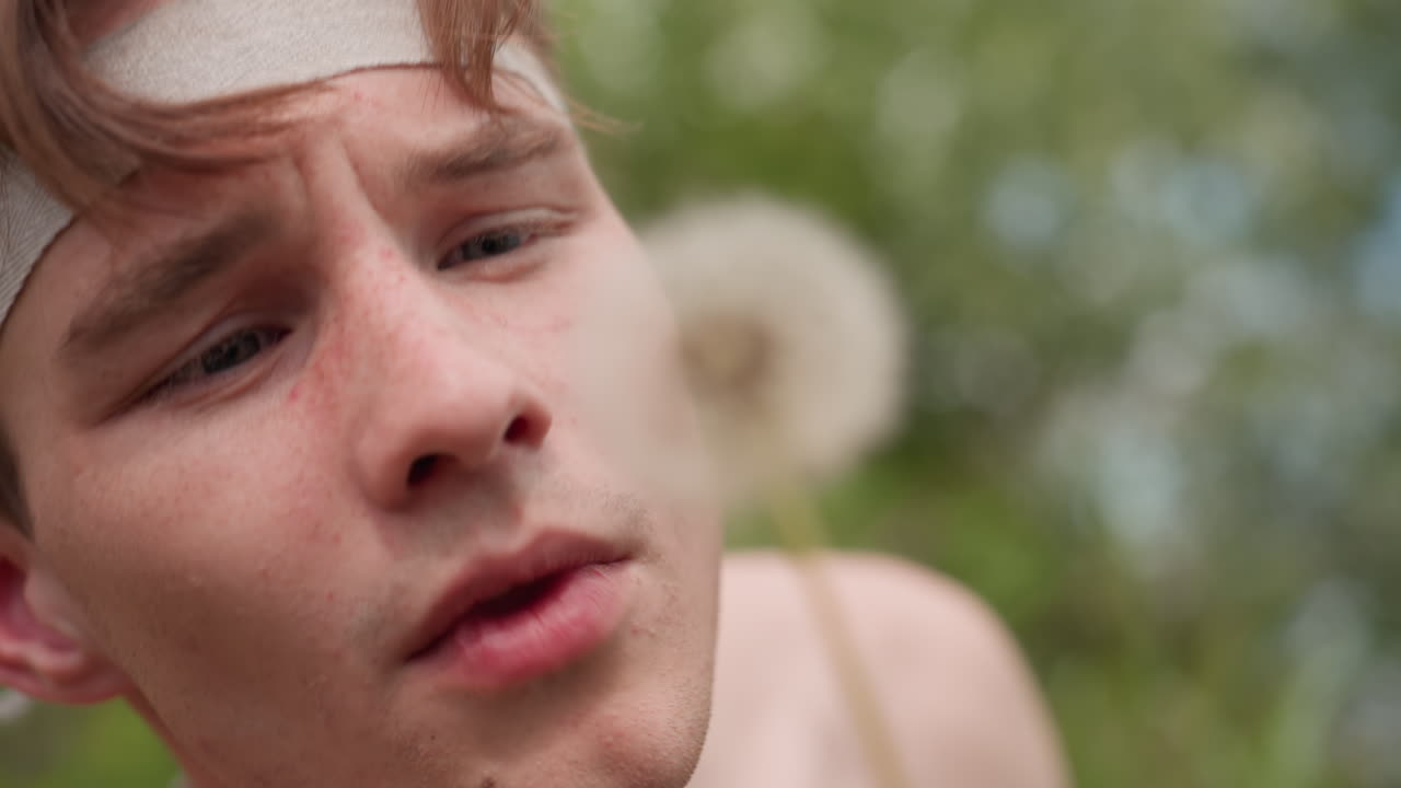 Close view of young man with brown hair, light freckles, and focused gaze holding dandelion near face, about to blow its seeds, surrounded by blurred greenery and soft natural scene