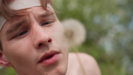 close view of young man with brown hair, light freckles, and focused gaze holding dandelion near face, about to blow its seeds, surrounded by blurred greenery and soft natural scene