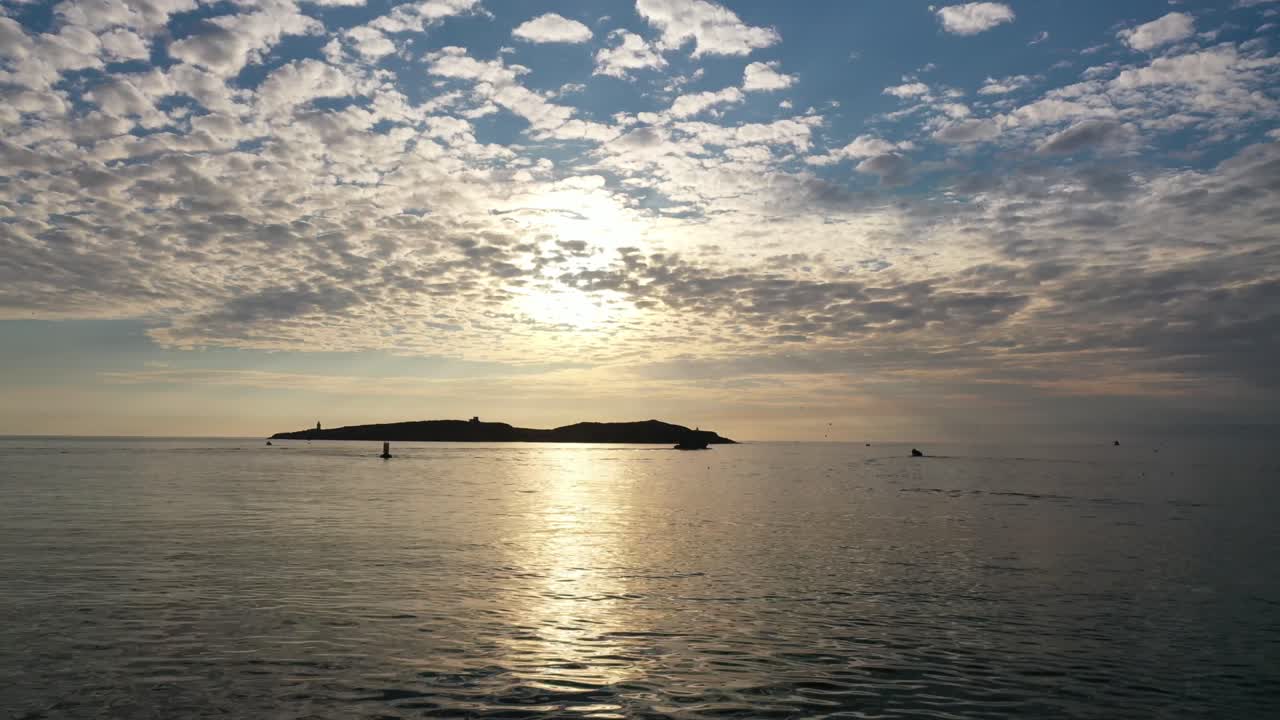 Cinematic drone shot orbiting over the Atlantic Ocean, circling around the dark silhouette of Mogador Island under a textured mackerel sky during golden hour