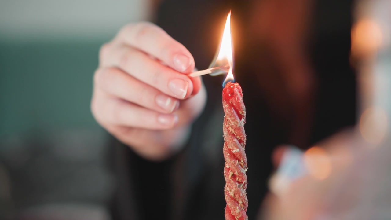 Mano che accende una candela attorcigliata con un fiammifero, sfondo bokeh sfocato, bagliore di lenta combustione, primo piano dello stoppino e della cera, donna che esegue un intimo rituale di divinazione, luce ambientale suggestiva che evoca la predizione.