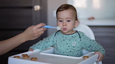 caring mother is feeding her baby boy from a spoon. mom's hand giving a spoon to her kid sitting in a feeding chair. blurred backdrop.