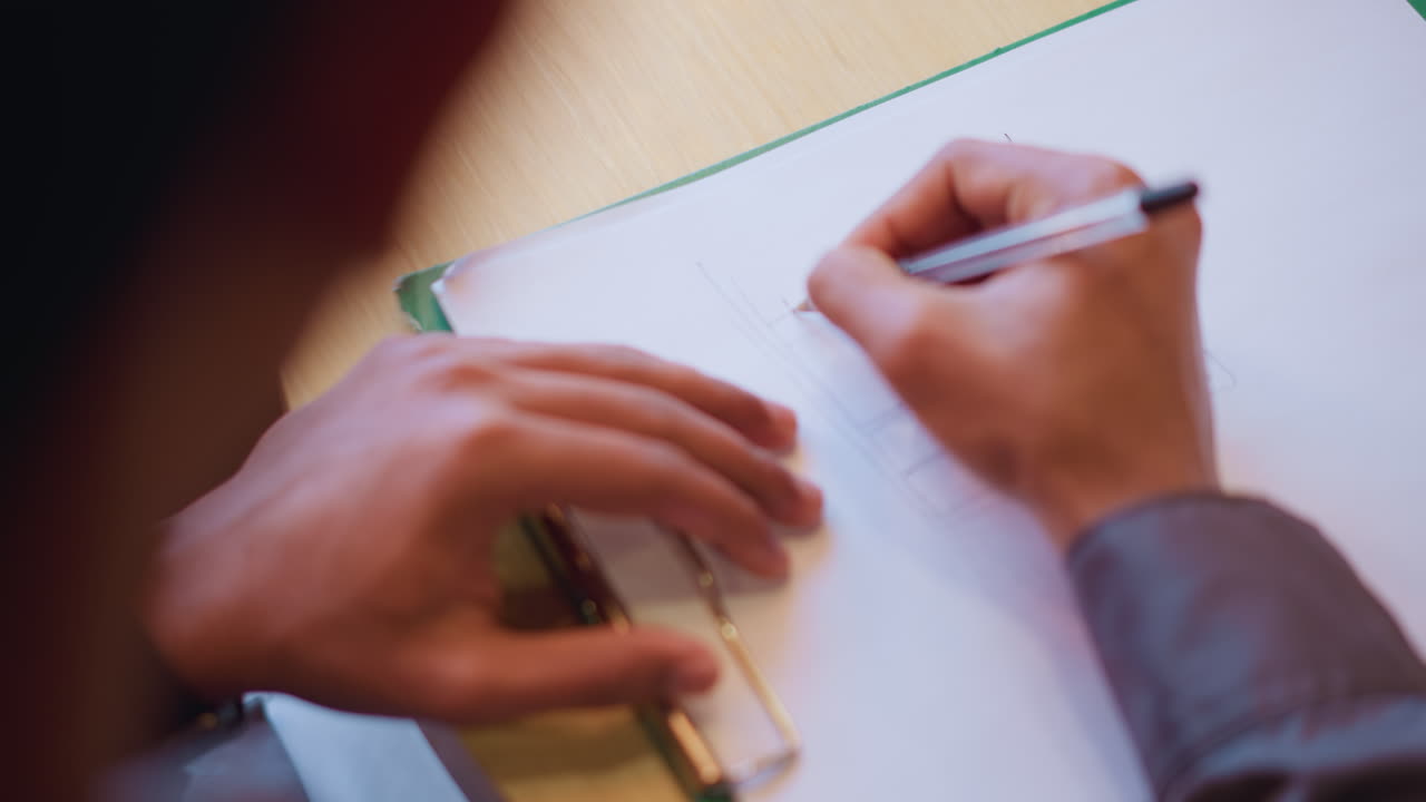 Person actively sketching technical design using black pen on clean sheet clipped to folder, viewed from above with focused hand gestures, demonstrating conceptual thinking during creative design process