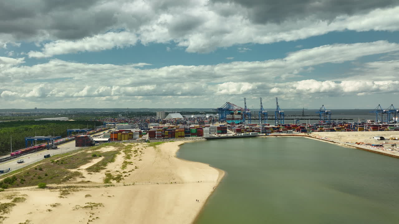 Aerial view of the Port of Gdańsk and Stogi Beach, featuring industrial cranes, shipping containers, and a sandy coastline under a partly cloudy sky