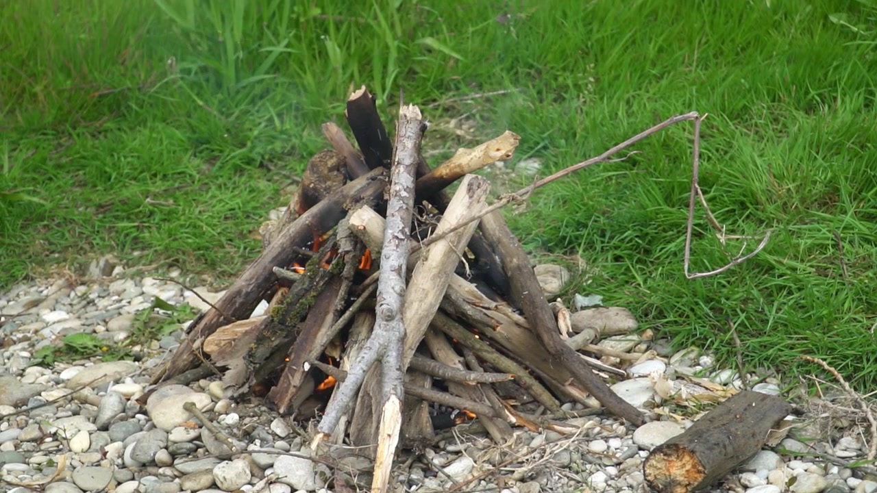 Szczegółowy widok nieoświetlonego ogniska w kształcie tipi zbudowanego z suchego drewna na kamieniach, otoczonego bujną trawą. Idealne do biwakowania na świeżym powietrzu lub motywów survivalowych.