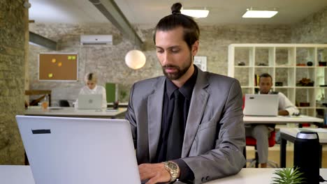Handsome businesswoman with beard is typing on laptop in office, watching at camera, smiling, colleagues are networking with technologies, work concept, communication concept