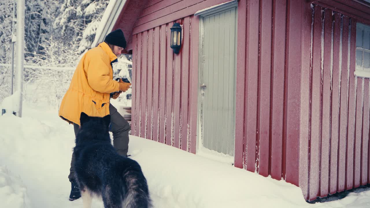 hombre caucásico con mascota de perro malamute de alaska entrando en una cabaña durante el invierno