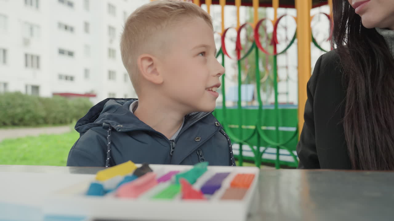 Escena de mesa al aire libre con un niño blanco sonriendo con confianza mientras discute planes escolares con su cuidador. Crayones y un cuaderno de trabajo en la mesa, contacto visual juguetón, postura relajada, un adulto de apoyo escuchando, en un vecindario.