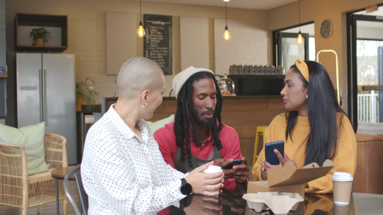Server and two customers greeting at cafe, leaning at table, viewing smartphone screen and laughing