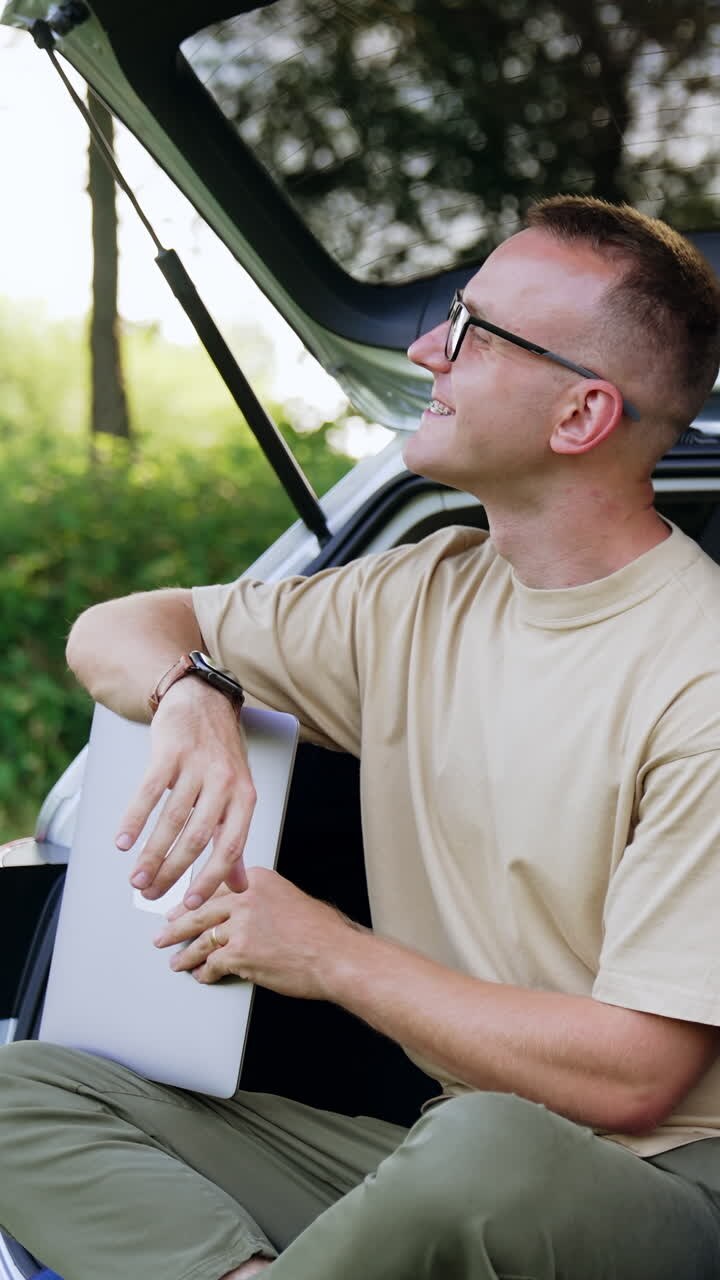 Uomo caucasico sorridente seduto nel bagagliaio dell'auto. L'uomo ha finito di lavorare al laptop ed è felice. Natura sullo sfondo. Video verticale.