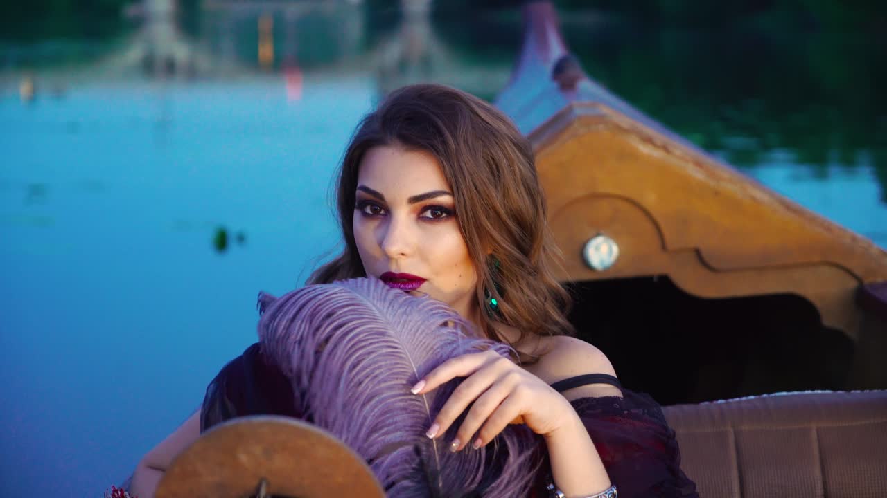 A beautiful woman playfully is glancing from under the guise of a purple feather on the gondola on a warm evening. Blurred background. Close-up