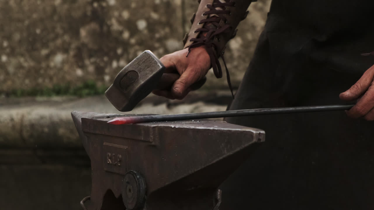 Static close-up of a blacksmith hammering a glowing iron rod on an anvil; metal flakes shear off with each strike as the tip is shaped to form