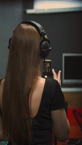 Elegant woman singer with headphones in black dress with open back performs song with microphone dancing in recording studio close backside view