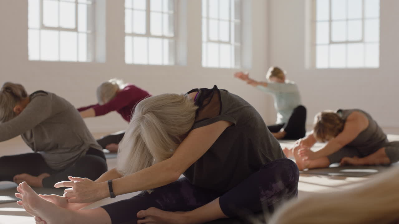 Yogaklasse med en sund moden kvinde, der praktiserer hoved-til-knæ foroverbøjning og nyder en fysisk morgentræning i et studie