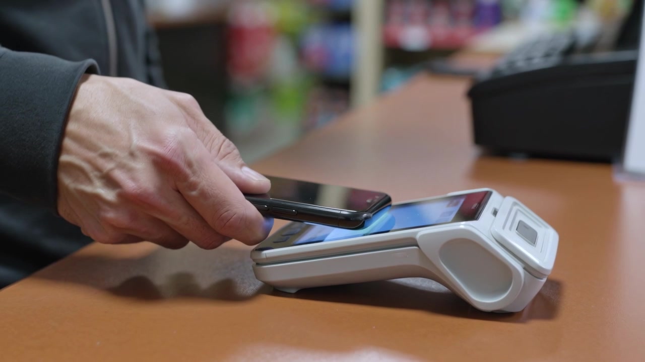 Starting NFC payment, man lowering phone over shop counter holding near white reader in dark hoodie