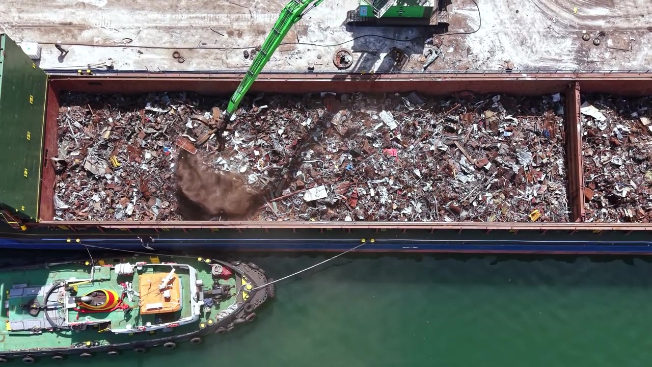 Rottami metallici vengono caricati su una nave da carico in un porto, sotto la luce del sole pomeridiano, utilizzando una gru e un rimorchiatore per il trasporto verso un'altra località per il riciclo o la lavorazione.