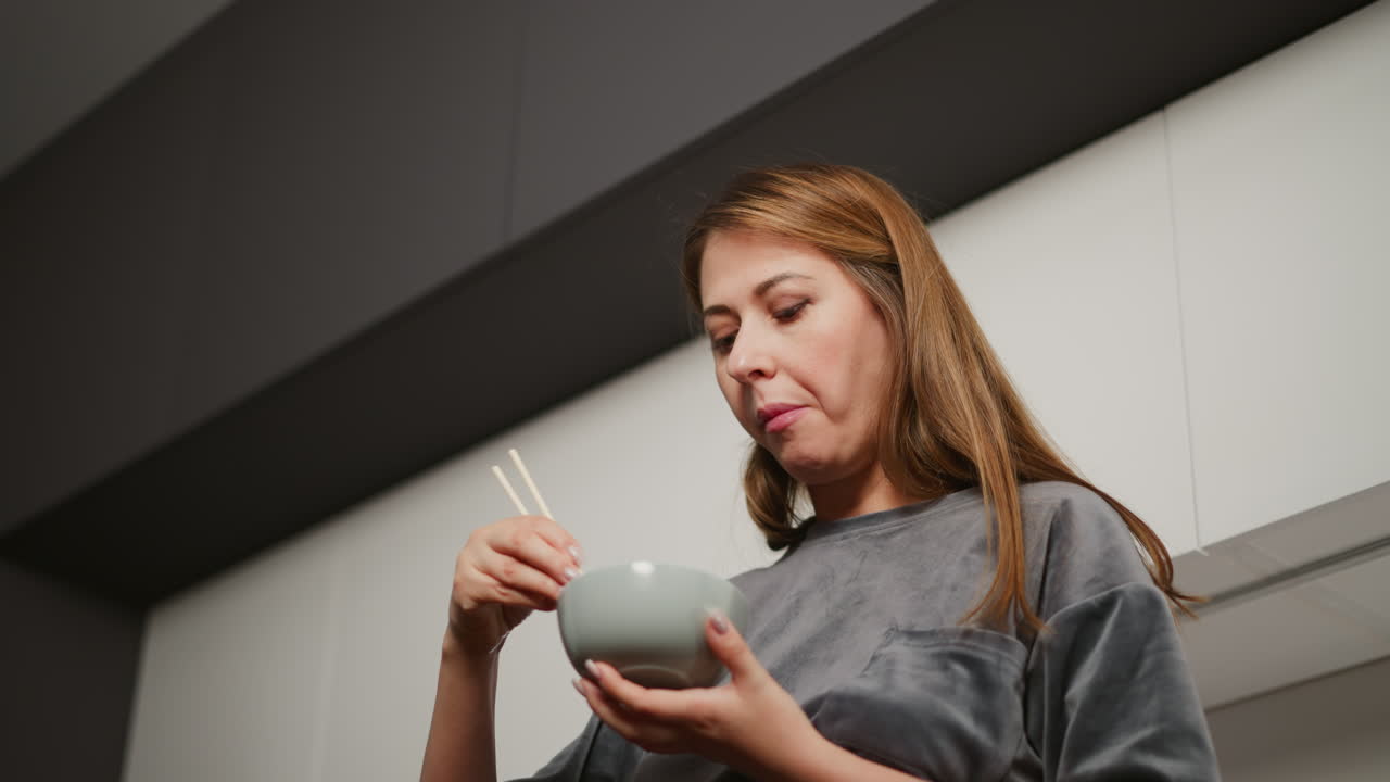 Upward angle shows woman in ash gray top holding bowl and chopsticks while eating breakfast in modern kitchen with sleek cabinetry and under-cabinet lighting