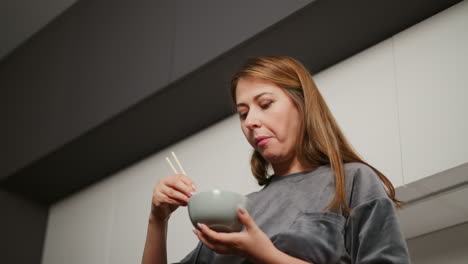 upward angle shows woman in ash gray top holding bowl and chopsticks while eating breakfast in modern kitchen with sleek cabinetry and under-cabinet lighting