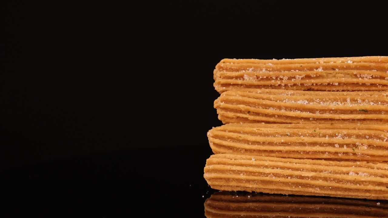 A neatly stacked pile of sugar-coated churros rotates smoothly on a glossy black surface, illuminated by soft studio lighting against a seamless dark backdrop
