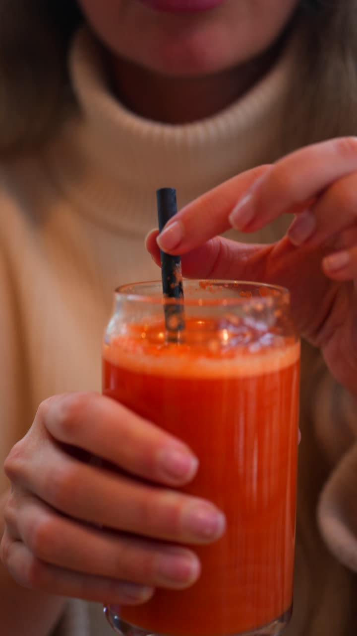 Primo piano di una donna che beve un succo di carota e arancia in un bicchiere con una cannuccia nera su un tavolo di legno. Verticale