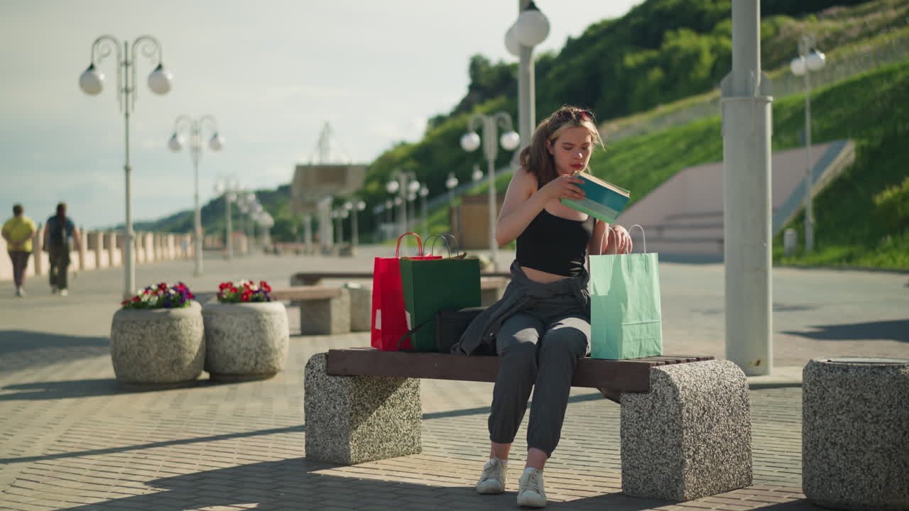 두 쪽 모두에 쇼핑 가방이 있는 야외 벤치에 앉아있는 여인이 민트 녹색 가방에서 책을 가져와 갈색 책을 같은 가방으로 돌려줍니다.