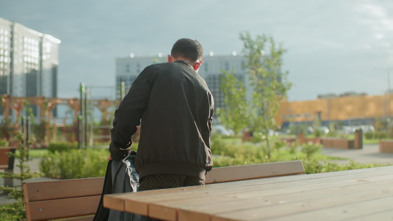Boy standing outdoors drops backpack on bench and begins to unzip, sunlight shining brightly in background with park greenery and blurred urban buildings