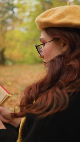 mujer con boina amarilla con el cabello rojo en cascada sobre el hombro lee un libro sola en el tranquilo bosque de otoño. inmersa en el pensamiento, hojea las páginas, disfrutando de la tranquila soledad