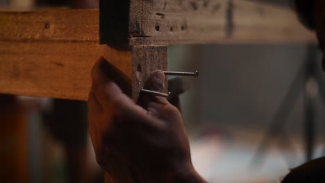 closeup of a male carpenter using a hammer to hammer the nails in a wooden plank, carpenter building furniture for home in his workshop