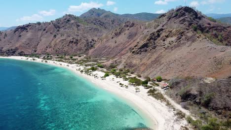 Aerial-drone-of-beautiful-white-sandy-beach,-crystal-clear-turquoise-ocean-water-and-rugged,-dry-and-arid-hill-tropical-island-landscape-of-capital-Dili,-Timor-Leste,-Southeast-Asia