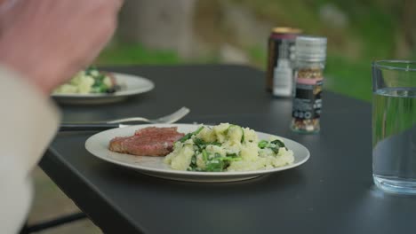 Person-seasoning-mashed-potatoes-and-steak-on-an-outdoor-table