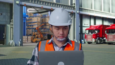 close up of asian male engineer with safety helmet working on a laptop while standing , outside of logistics distributions warehouse
