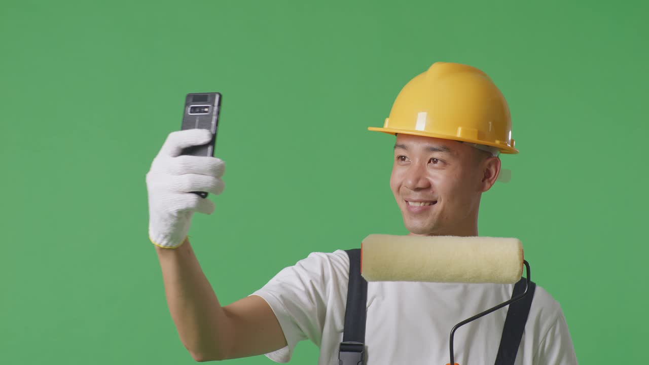 Close Up Of Asian Man Painter Wearing Safety Helmet Using Smartphone Taking Picture While Standing In The Green Screen Background Studio