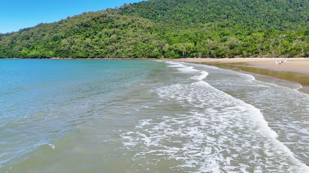 Un dron se desliza sobre las tranquilas olas, la playa de arena y la exuberante selva tropical bajo la brillante luz del día