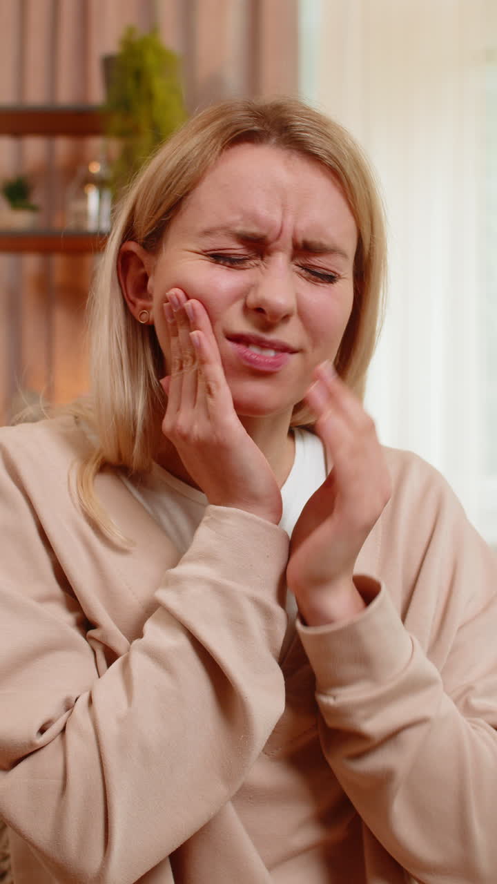 Adult woman sitting on sofa holding cheek with hand showing strong tooth pain discomfort at home