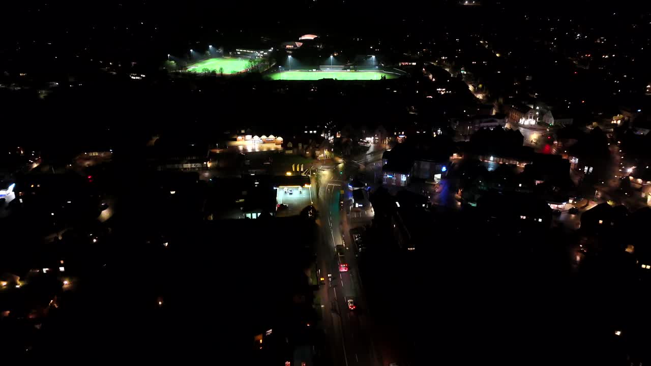 Small american town with driving cars on lighting street. Night scene with illuminated soccer fields in background. Aerial forward wide shot.