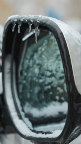 close-up of a car mirror covered in snow with icicles with reflection of the car window , showing a blurred background of another snow-covered car in winter