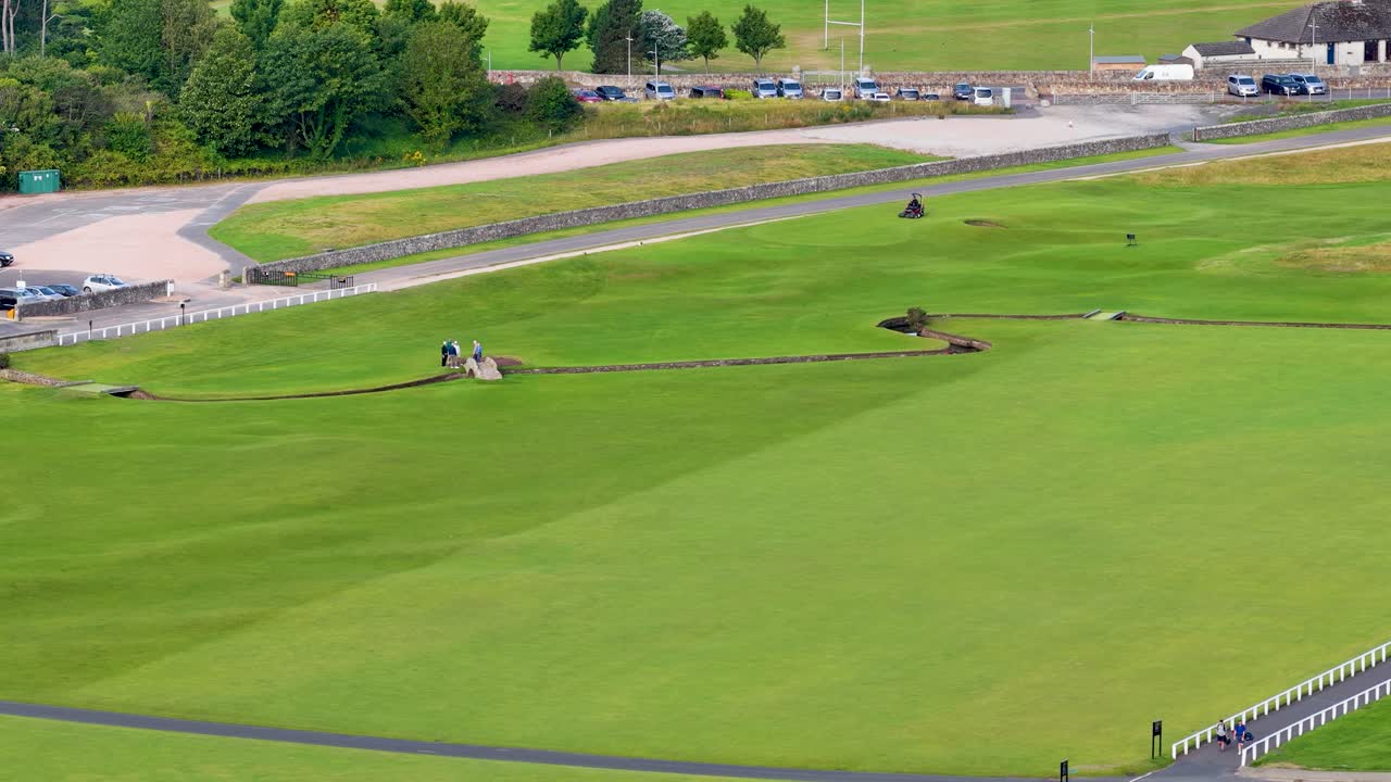 Golfers cross iconic stone bridge on lush fairway, wide aerial view, bright natural daylight