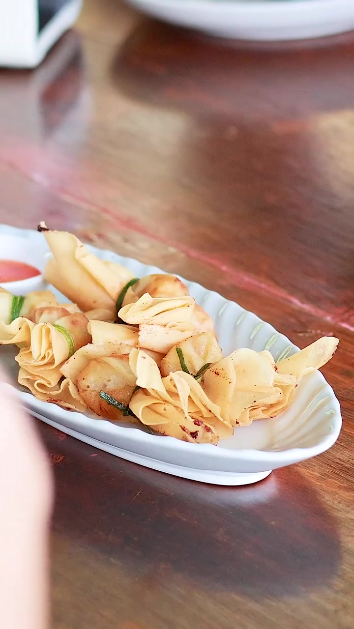 Una persona serve ravioli a sacchetto tailandesi su un tavolo di legno, evidenziando un ambiente da pranzo informale con illuminazione naturale.