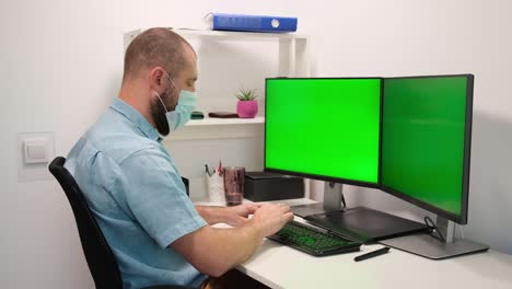 hombre con máscara médica escribiendo en el teclado frente a dos monitores verdes. trabajo remoto en casa en cuarentena