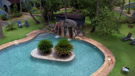 resort swimming pool with waterfall surrounded by lush vegetation, aerial orbit shot