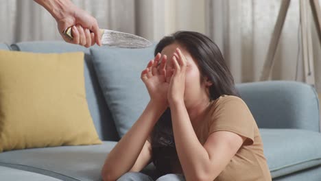 close up of young asian woman victim of violence with bruise on body sits on the floor being scared of a man holding a knife threatening her at home