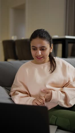 Vídeo-Vertical-De-Una-Chica-Morena-Feliz-Con-Pantalones-De-Color-Rosa-Claro-Comunicándose-Por-Videoconferencia-Usando-Una-Computadora-Portátil-En-Un-Apartamento-Moderno