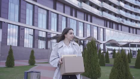 a happy woman is walking in front of an office building. she carries her things in a box. she has quitted her job. she is smiling.