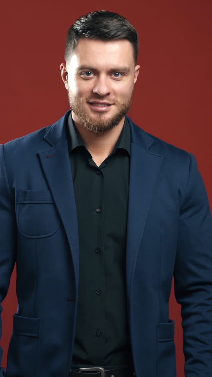 Handsome man in stylish suit. Portrait of young businessman in the red studio. Smiling bearded man in black shirt and blue jacket looking at camera. Vertical video