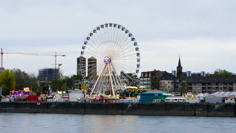 wide panning handheld shot of a moving ferris wheel in cologne germany glowing in different colors at a festival with other attractions and rides during dusk